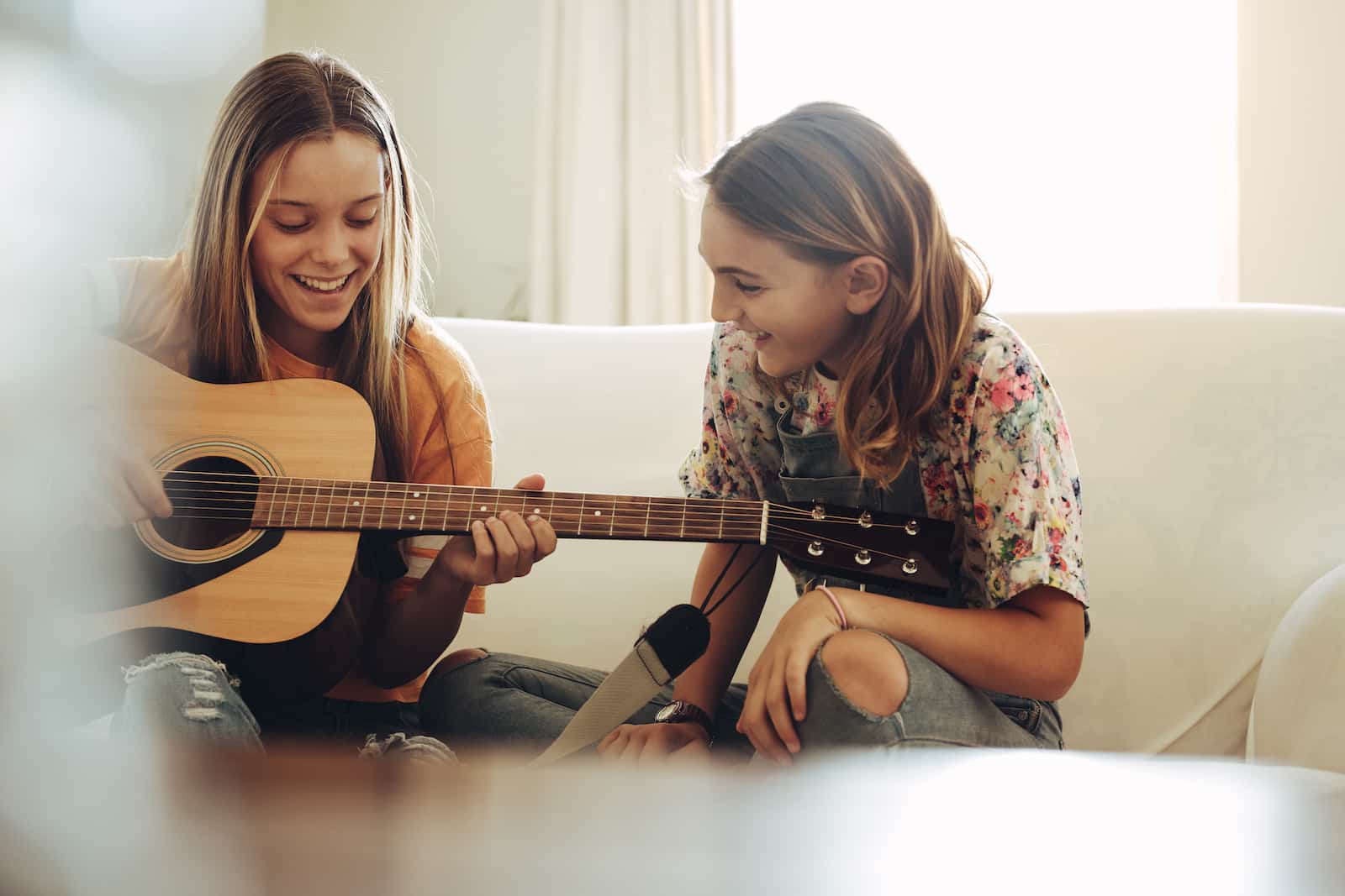 Two teenage girls sitting together at home and having fun playing guitar. Girl learning to play guitar at home sitting with her friend.